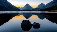 Tranquil mountain lake at sunrise with perfect reflection of peaks in calm water, morning mist hovering over surface, two rocks emerging from glassy waters.