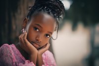 Pensive young African girl with braided hair in pink dress resting chin on hand, soft bokeh background creates dreamy atmosphere for childhood portraits.
