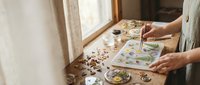 Woman artist painting watercolor botanical illustrations at wooden table by window with natural light and art supplies scattered around workspace.