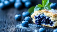Fresh blueberry pastry with powdered sugar and mint garnish on dark wooden background, macro shot showing flaky layers and juicy fruit filling.