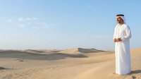 Middle Eastern man wearing traditional white thobe and ghutra headwear standing in vast desert landscape with sand dunes under clear blue sky, suitable for cultural, travel, and lifestyle content.
