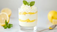 Layered lemon dessert with whipped cream and mint garnish in glass, creamy vanilla mousse and citrus curd, fresh ingredients on light background.