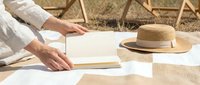 Woman in white linen clothing reading book outdoors on sunny day with straw hat nearby, relaxing summer vacation lifestyle concept for wellness and leisure.