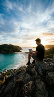 Young man working on laptop while sitting on rocky cliff overlooking turquoise ocean bay at sunset for remote work and digital nomad lifestyle.