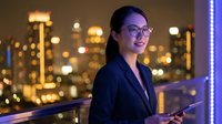 Young Asian businesswoman in glasses holding tablet on rooftop terrace with glowing city skyline at night, confident professional smile.