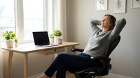 Young professional woman relaxing in ergonomic office chair at home workspace with laptop, taking break from remote work in bright modern interior.