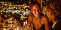 Young blonde woman laughing during romantic dinner date with wine glasses and city lights bokeh background creating warm intimate atmosphere for couples dining.