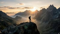 Lone hiker silhouette standing on rocky cliff edge overlooking misty mountain valley at golden sunrise with dramatic peaks and layered ridges.