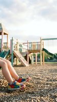 Child's legs in colorful sneakers resting on wood chip ground at a sunny playground with slide and climbing structure in background.