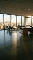 Modern high-rise office space with floor-to-ceiling windows overlooking city skyline, featuring wooden desk, black chairs, and polished concrete flooring for corporate workspace design.