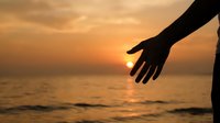 Hand reaching out during golden hour sunset over ocean waves creating warm romantic atmosphere for connection and support concepts.