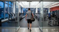 Athletic man entering modern fitness center with gym bag, walking through glass doors toward cardio and weight training equipment in spacious health club facility.