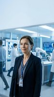 Middle-aged woman with dark hair wearing navy blazer and ID badge stands in modern laboratory facility with glass partitions, medical equipment, and colleagues working in background.
