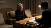 Senior man with gray hair and mature Caucasian appearance in business meeting with younger man, both wearing dark suits at wooden table in office setting for corporate negotiation or consultation.