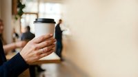 Person holding takeaway coffee cup with lid in office environment, suitable for business, lifestyle, and beverage marketing materials.