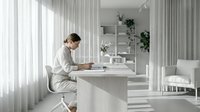 Young professional woman working on laptop in modern minimalist white kitchen with natural light streaming through curtains.