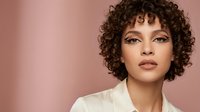 Professional portrait of confident mixed-race woman with curly hair and natural makeup against soft pink background for beauty and fashion campaigns.