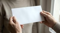 Mature adult male hands holding blank white paper card against neutral beige background, suitable for mockups, presentations, and business communications.