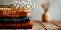 Stack of cozy knitted sweaters in autumn colors with dried flowers on wooden surface creating warm seasonal home decor atmosphere for fashion styling.