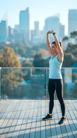 Woman in light blue tank top and black leggings stretching arms overhead on rooftop terrace with city skyline backdrop for fitness and wellness content.