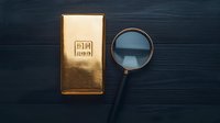 Gold bar with 999 purity mark and magnifying glass on dark wooden surface, symbolizing precious metal investment analysis and authenticity verification.