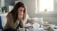 Young woman with long brown hair blowing nose with tissue while sitting on couch at home, feeling unwell with cold or flu symptoms in bright living room.