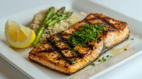 Grilled salmon fillet with fresh herbs, asparagus and quinoa on white plate. Charred fish garnished with parsley and lemon wedge, healthy protein dinner.