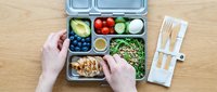 Person with light skin tone preparing healthy meal prep lunch box with fresh vegetables, fruits, grains and protein on wooden surface for diet planning and nutrition.