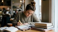 Young woman student writing notes in notebook while studying at cafe table with books and coffee cup for academic research and learning.