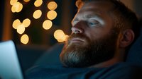 Caucasian bearded man working late on laptop with Christmas lights bokeh in background creating cozy atmosphere for remote work from home office setting.