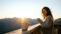 Woman with long dark hair, South Asian ethnicity, wearing a cozy cardigan, holding a warm beverage while enjoying sunrise over mountains from a wooden deck at dawn.