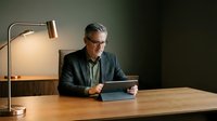 Middle-aged man with gray hair and glasses, wearing dark blazer and light shirt, sitting at wooden desk using tablet device under warm desk lamp for work or study.
