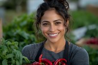 Young Hispanic woman farmer with bright smile holding fresh red bell peppers in garden setting, wearing casual gray top with natural curly updo hairstyle.