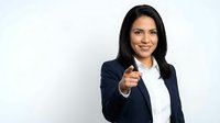 Confident young Hispanic businesswoman in navy suit pointing at camera with a smile, isolated on light gray background.