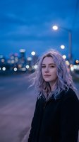 Young blonde woman in black jacket standing on urban street at twilight with city lights and bokeh background creating atmospheric evening mood.