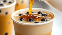 Pouring golden caramel syrup into creamy bubble milk tea with black tapioca pearls, close-up view showing liquid stream and boba texture in plastic cup.