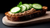 Fresh cucumber slices on dark rye bread with liver pate spread, served on wooden cutting board against black background. Food photography.
