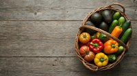 Wicker basket filled with fresh organic vegetables including peppers, tomatoes, and cucumbers arranged on rustic wooden surface for farmers market, grocery, or healthy eating concepts.
