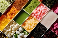 Colorful assortment of tea ingredients and toppings in compartment box including matcha powder, tapioca pearls, dried flowers, and fruit jellies for bubble tea preparation.