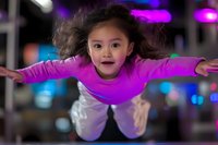 Young Asian girl in pink top and white pants flying through air with arms spread wide, expressing joy and freedom against colorful bokeh background.