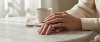 Woman hands with wedding ring resting on marble table next to coffee cup in soft morning light creating peaceful domestic moment.