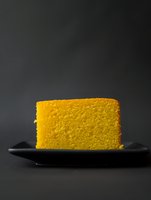 Slice of golden sponge cake on black plate against dark background, showcasing soft texture and classic baking. Minimalist food photography for restaurant menus.