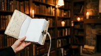 Person holding open blank notebook with beige textured cover in library setting with wooden bookshelves and warm lighting for journaling, planning, writing projects.