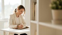 Professional businesswoman in beige blazer writing notes at modern white desk in bright office interior with natural lighting for corporate presentations.