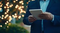 Businessman in navy suit using digital tablet with festive bokeh lights in background, creating professional holiday atmosphere for business content.