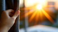 Hand pulling back curtain revealing bright sunburst through window at sunrise, creating atmospheric morning light and shadow play with fabric texture.