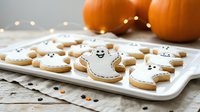 Decorated ghost-shaped cookies with white icing and smiling faces arranged on a white plate beside orange pumpkins and scattered candy for autumn holiday baking and festive celebrations.