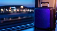 Purple travel suitcase on train window ledge with blurred night city lights in background, creating atmospheric travel mood.