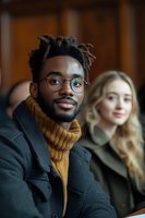 Young African American man with glasses and mustard turtleneck sweater smiling at camera, wearing winter coat. Blonde woman blurred in background. Professional portrait.
