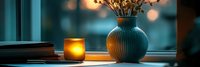 Cozy evening ambiance with glowing candle and dried flowers in ceramic vase on windowsill against blurred city lights background.